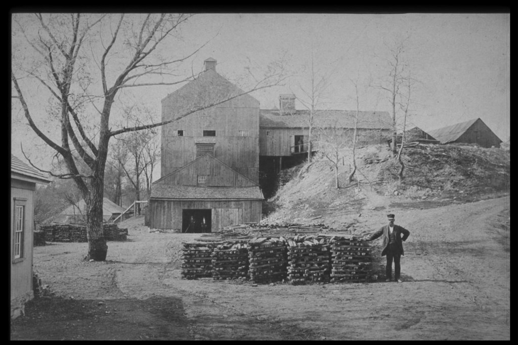 The Sharon Valley Iron Company furnace in 1875. Note the stacks of pig iron in the foreground and beyond the tree to the left. To the upper right stand the charcoal sheds. At the same level is the elongated casting shed running into the main building to the top of the thirty-four-foot-high blast furnace. Above the unseen furnace is the top house extending sixty-eight feet above ground. To the far left is the blast house with an overshot waterwheel powering pumping tubs through the visible pipe to the top house to heat the blast. The casting shed, center front, contains the sand beds where the white-hot iron formed into pigs and cooled. - Salisbury Association