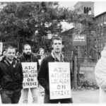 Black and white photo of people walking on a sidewalk with signs hung around their neck in front of a industrial building.