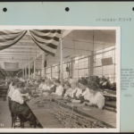 Black and white photograph of women sitting at long tables inspecting small parts of pistols. American flags drape from the ceiling. The photograph is mounted on a larger piece of paper.
